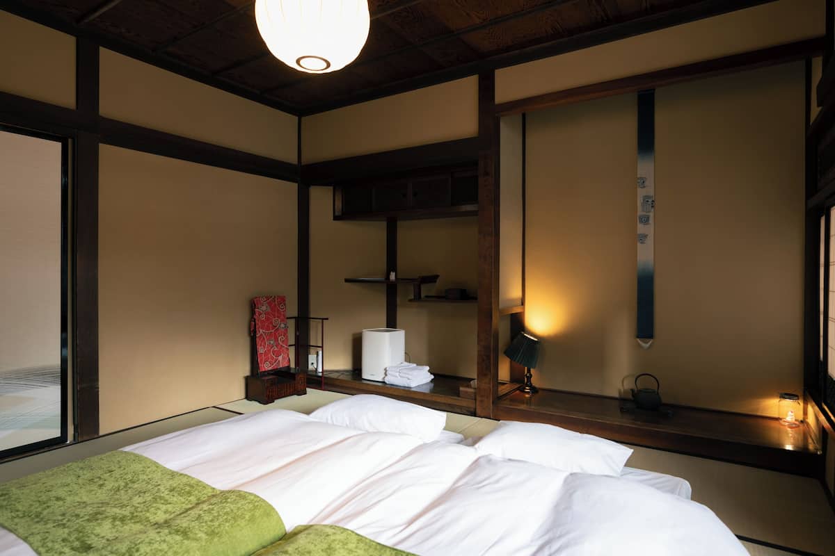 A traditional Japanese-style room with tatami flooring, a low wooden ceiling, and a large shoji screen. There is a bed with white bedding and a green throw blanket, a small table with a lamp, and a mirror on the wall.