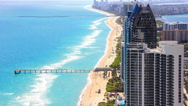 Un hotel moderno con vista a la playa, un muelle que se adentra en el océano y un horizonte urbano al fondo.