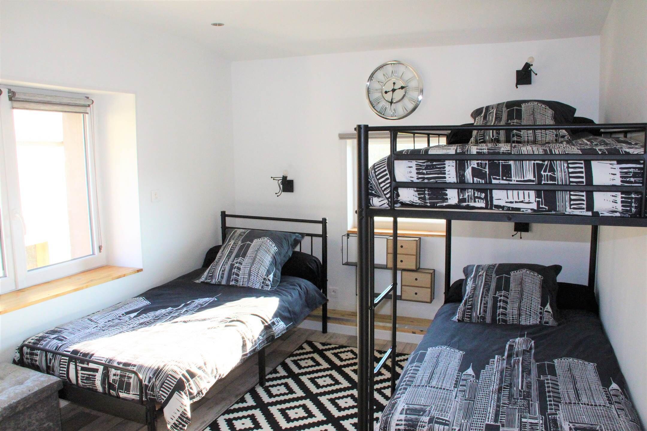 A dormitory room with bunk beds, a window, a clock, and a patterned rug.