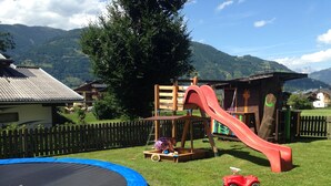 Ein Spielplatz mit Rutsche und Trampolin, umgeben von einem Holenzaun und Häusern in einer hügeligen Landschaft.