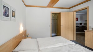Ein Schlafzimmer mit einem hölzernen Kopfteil, einem Bett mit weißer Bettwäsche und einem kleinen Nachttisch mit einer Vase. An der Wand hängen Bilder in Rahmen, und eine Tür führt zu einem Badezimmer und einer Küche.