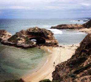 A coastal landscape with a natural rock formation featuring a cave entrance, a sandy beach, and the ocean.