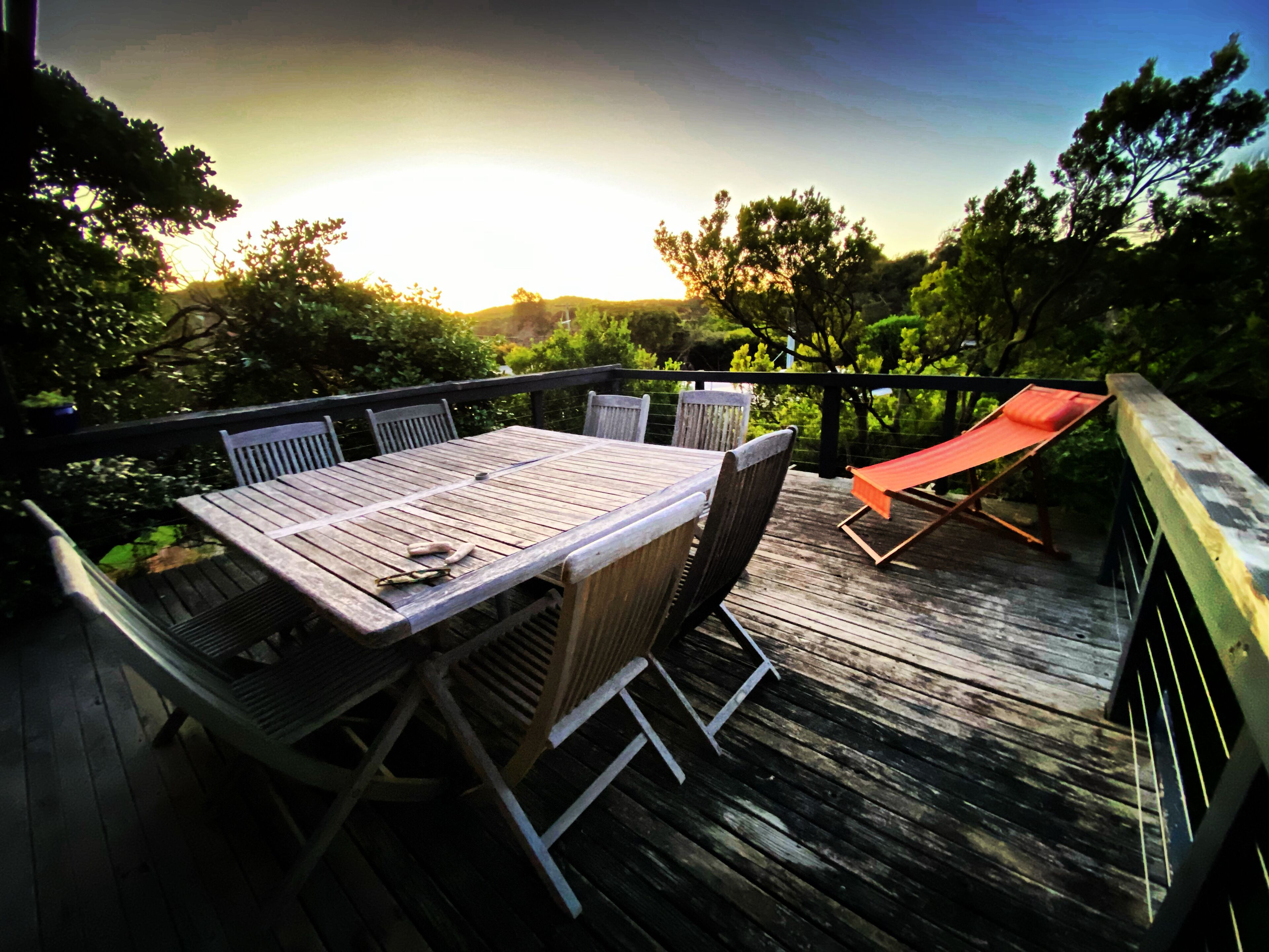 A wooden deck with a table and chairs, surrounded by trees and a sunset in the background.