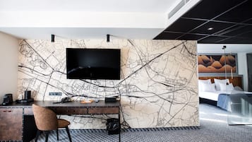 A hotel room with a large flat-screen TV mounted on a wall with a map design, a wooden desk with a chair, and a bed with a modern headboard.