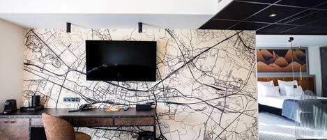 A hotel room with a large flat-screen TV mounted on a wall with a map design, a wooden desk with a chair, and a bed with a modern headboard.