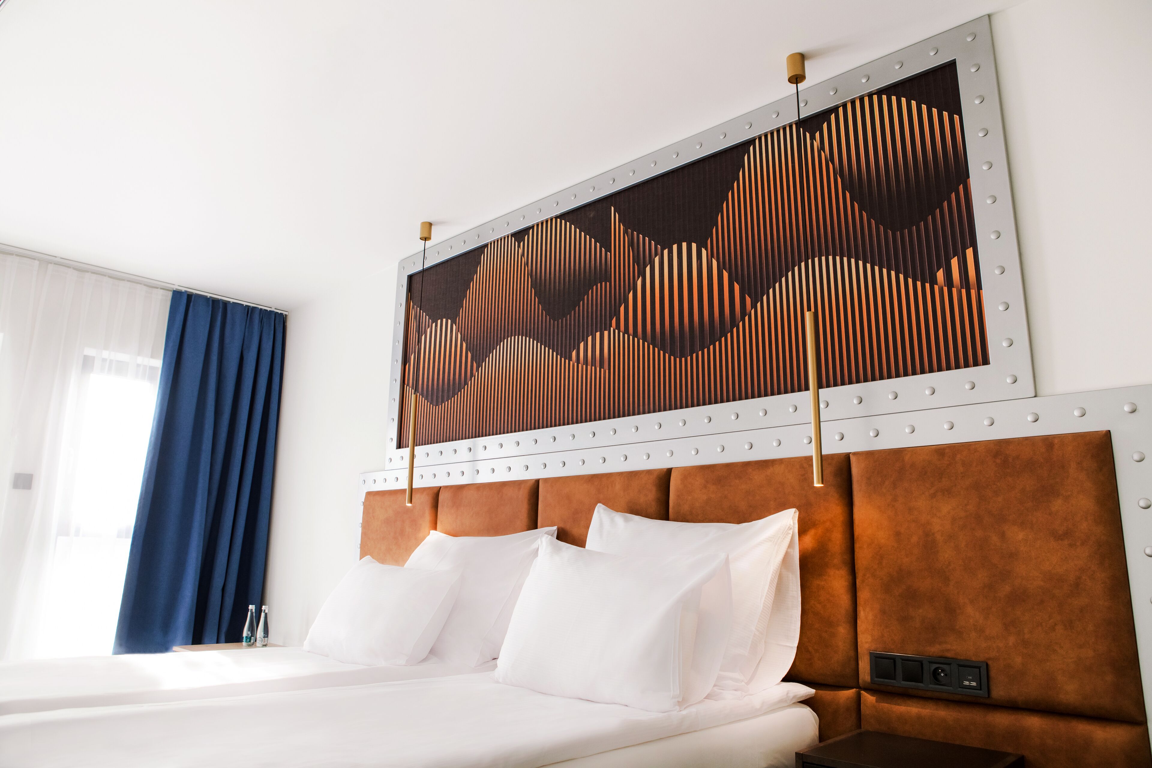 A hotel room with a large bed, a headboard with a geometric patterned design, and a window with blue curtains.