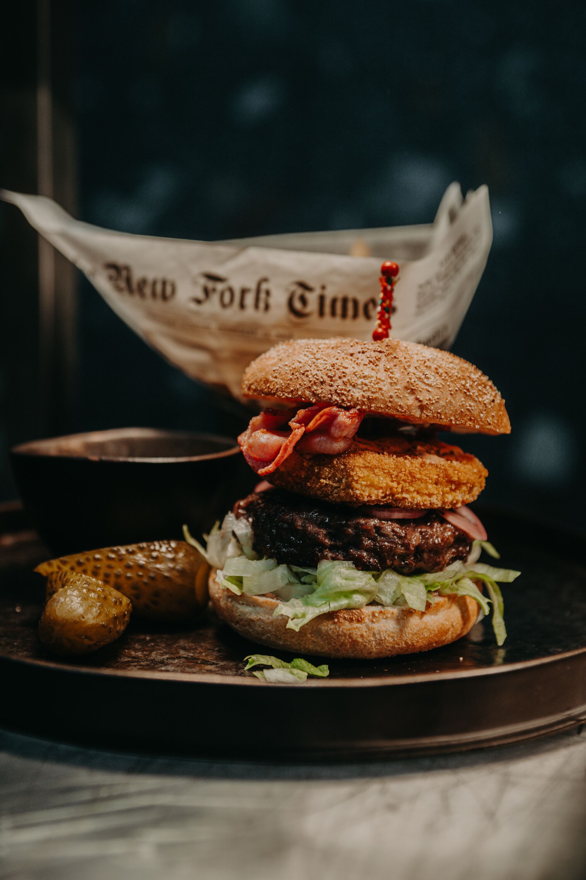 A burger with a sesame seed bun, bacon, and pickles on a plate.