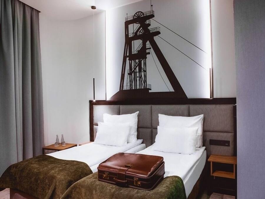 A hotel room with a large bed, two pillows, a brown suitcase, a nightstand, and a wall-mounted artwork of a mining structure.