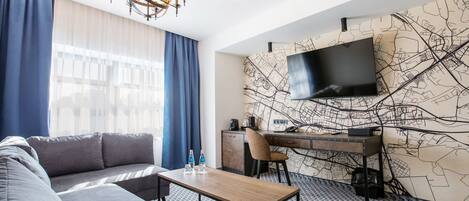 Une chambre d’hôtel moderne comprenant un canapé, une table basse en bois, un bureau avec une chaise, un téléviseur à écran plat fixé au mur et orné d’un motif représentant une carte de la ville, ainsi qu’un lustre.