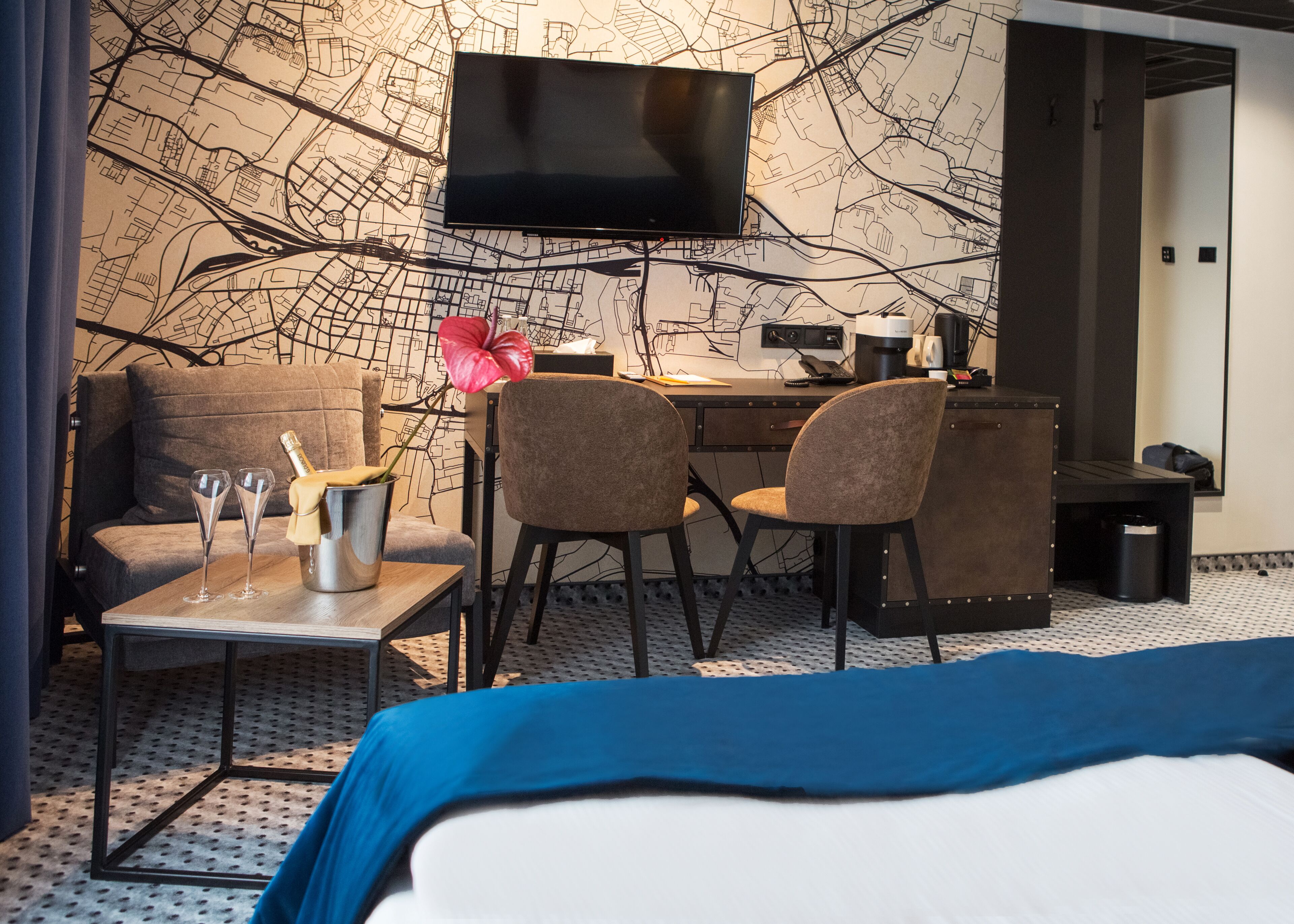 A hotel room with a bed, a desk, two chairs, a television, a map on the wall, and a small table with glasses and a bottle.