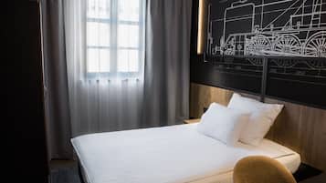 Habitación de hotel con una cama, cabecera de madera, un ventanal grande con cortinas traslúcidas y una obra de arte en la pared con motivo de un tren.