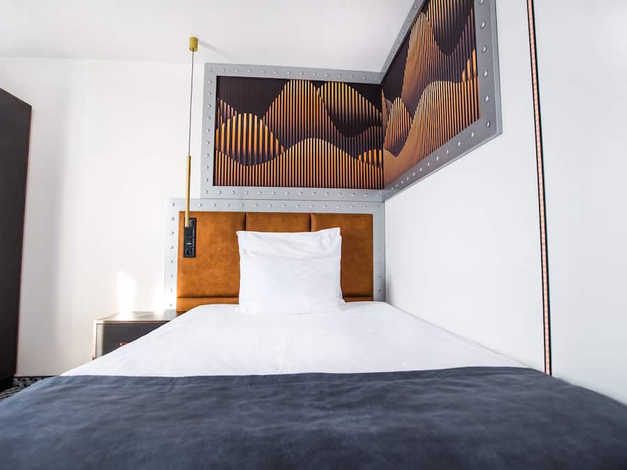 A hotel room with a bed, a nightstand, a wall art piece, and a wooden headboard.