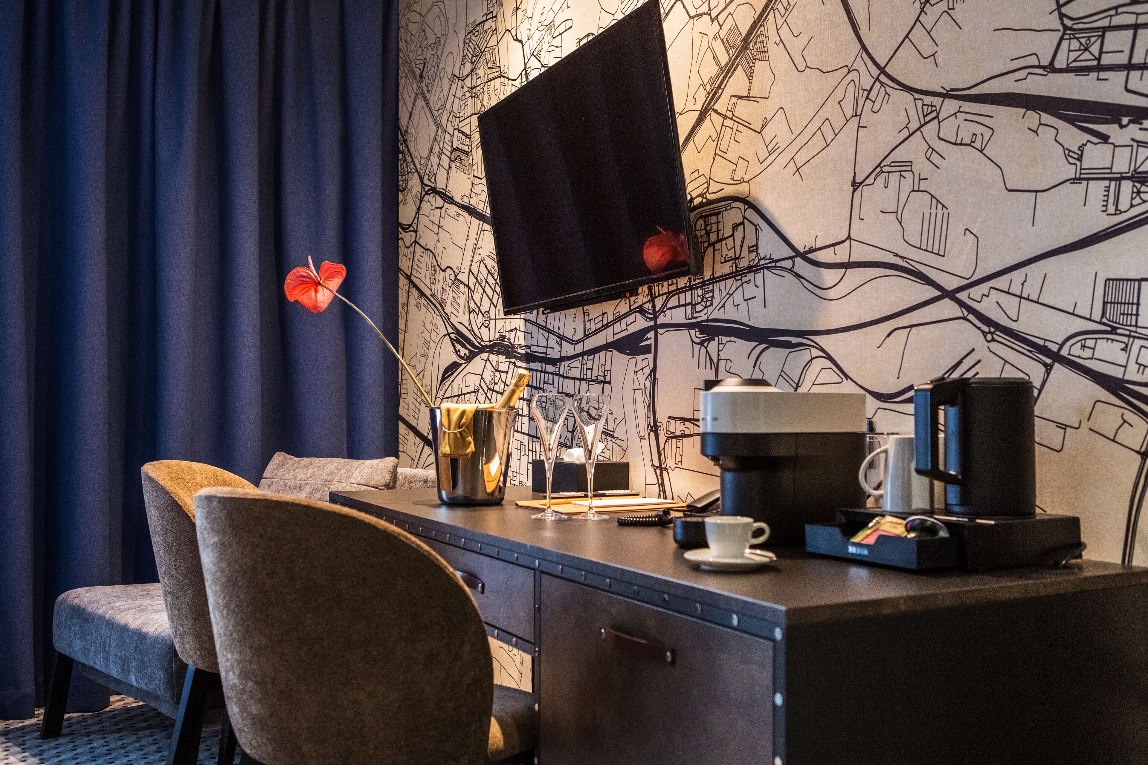 A hotel room with a desk, two chairs, a coffee maker, a cup, a vase with a red flower, and a map-themed wall.