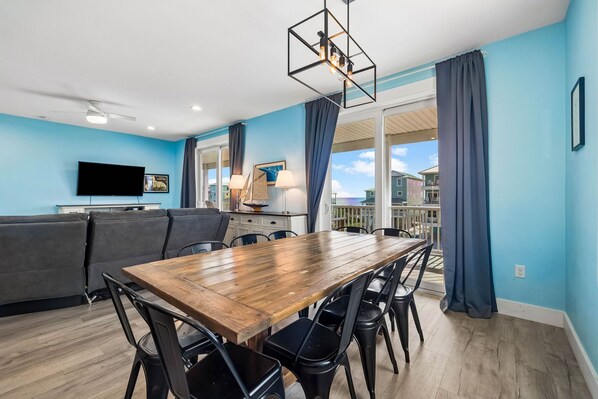 A modern living room with a wooden dining table, black chairs, a grey sofa, a flat-screen TV, and a ceiling fan.