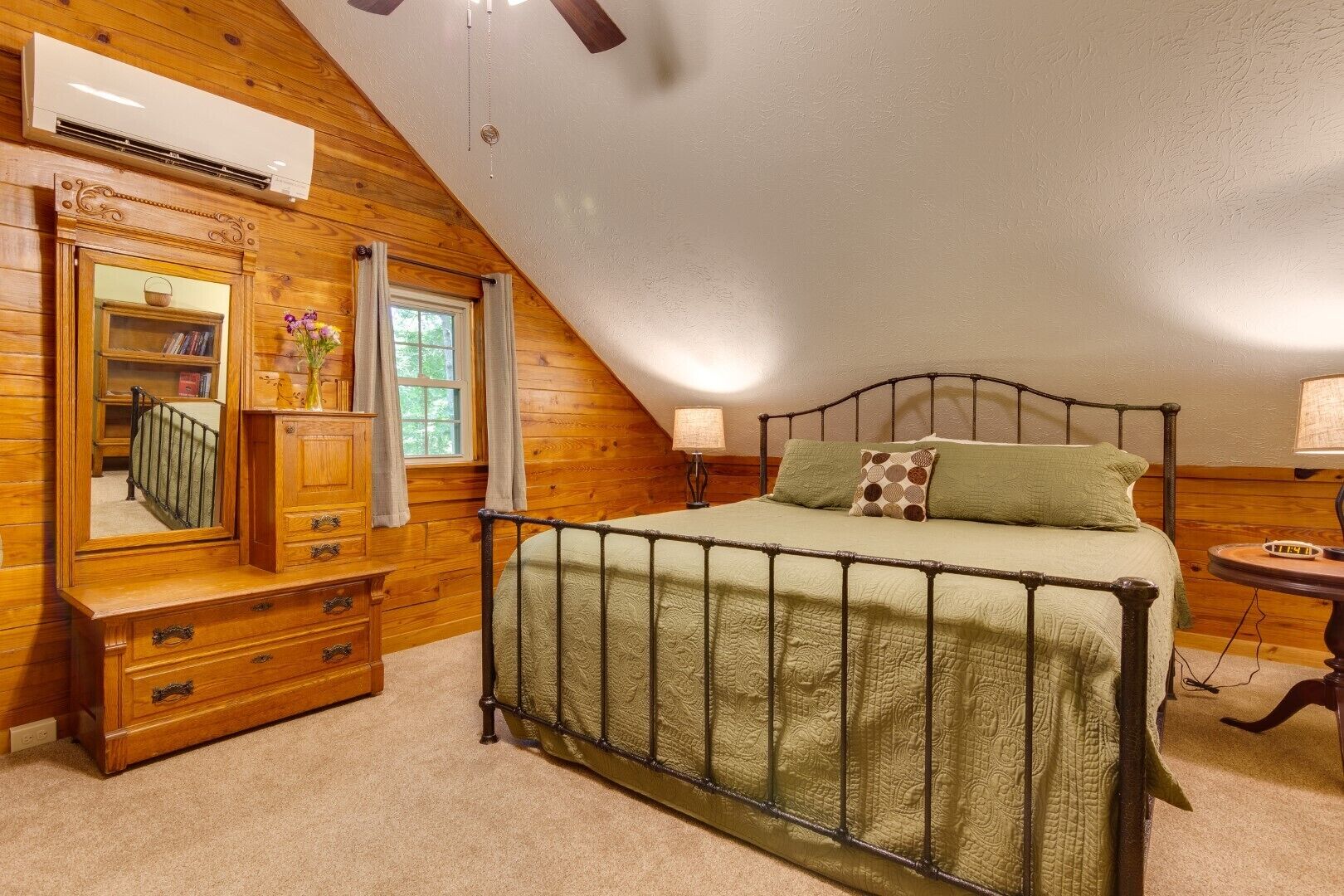 A bedroom with a sloped ceiling, a bed with a metal frame, a wooden dresser, a vanity with a mirror, and a window with curtains.