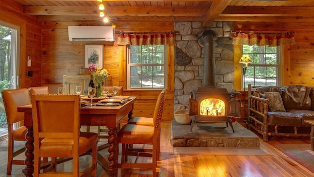 A cozy cabin interior with a stone fireplace, wooden walls, and a dining area with leather chairs.