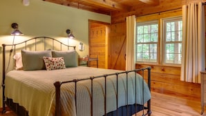 A wooden cabin bedroom with a bed, two wall-mounted lamps, a window with curtains, and a wooden door.
