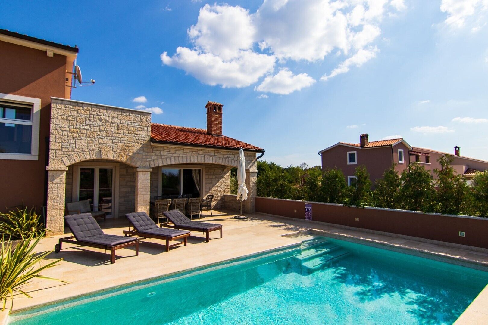 A swimming pool with lounge chairs, a stone building with a tiled roof, and a clear blue sky.