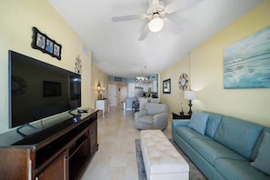 A living room with a TV, sofa, ottoman, ceiling fan, and artwork on the walls.
