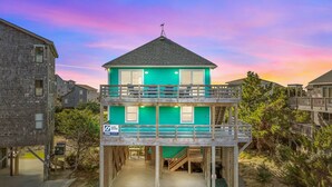 Une maison à deux étages avec un balcon à l’étage supérieur, un petit escalier menant à l’entrée et une enseigne indiquant un service de location d’équipements de surf.