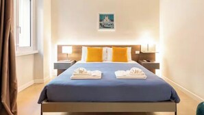 A hotel room with a bed, two bedside tables, a nightstand, a painting on the wall, and a window with curtains. - Largo Pannonia Home: A Gateway to Historic Rome Near the Colosseum and St. John & more (Roma)