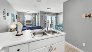 A modern kitchen with a white countertop, double sink, and a view of the living area with a blue sofa and curtains. - 🌴Cute 1BR|Steps2Beach w/ Incredible View|Pool!🌴 (Navarre Beach)