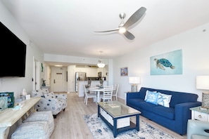 A modern living room with a blue sofa, a glass coffee table, and a ceiling fan. - Beautiful 9th Floor at The Palms of Destin - Hard Floors Throughout! (Destin)
