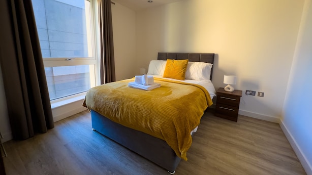 Quarto de hotel com cama, mesa de cabeceira, luminária e janela com cortinas.