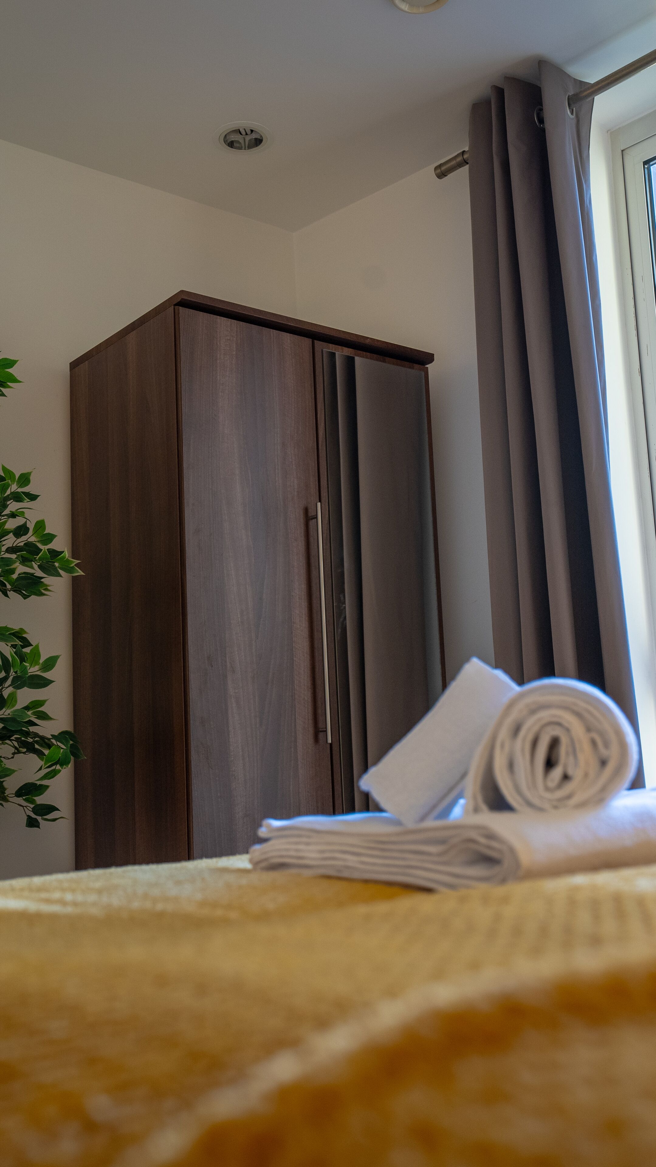 A bedroom with a wardrobe, a bed with a rolled-up towel, and a window with curtains.