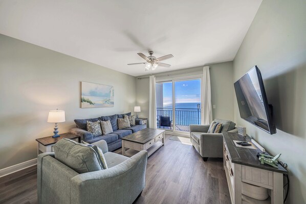 A spacious living room with a large window, a ceiling fan, a flat-screen TV, and a comfortable seating area.
