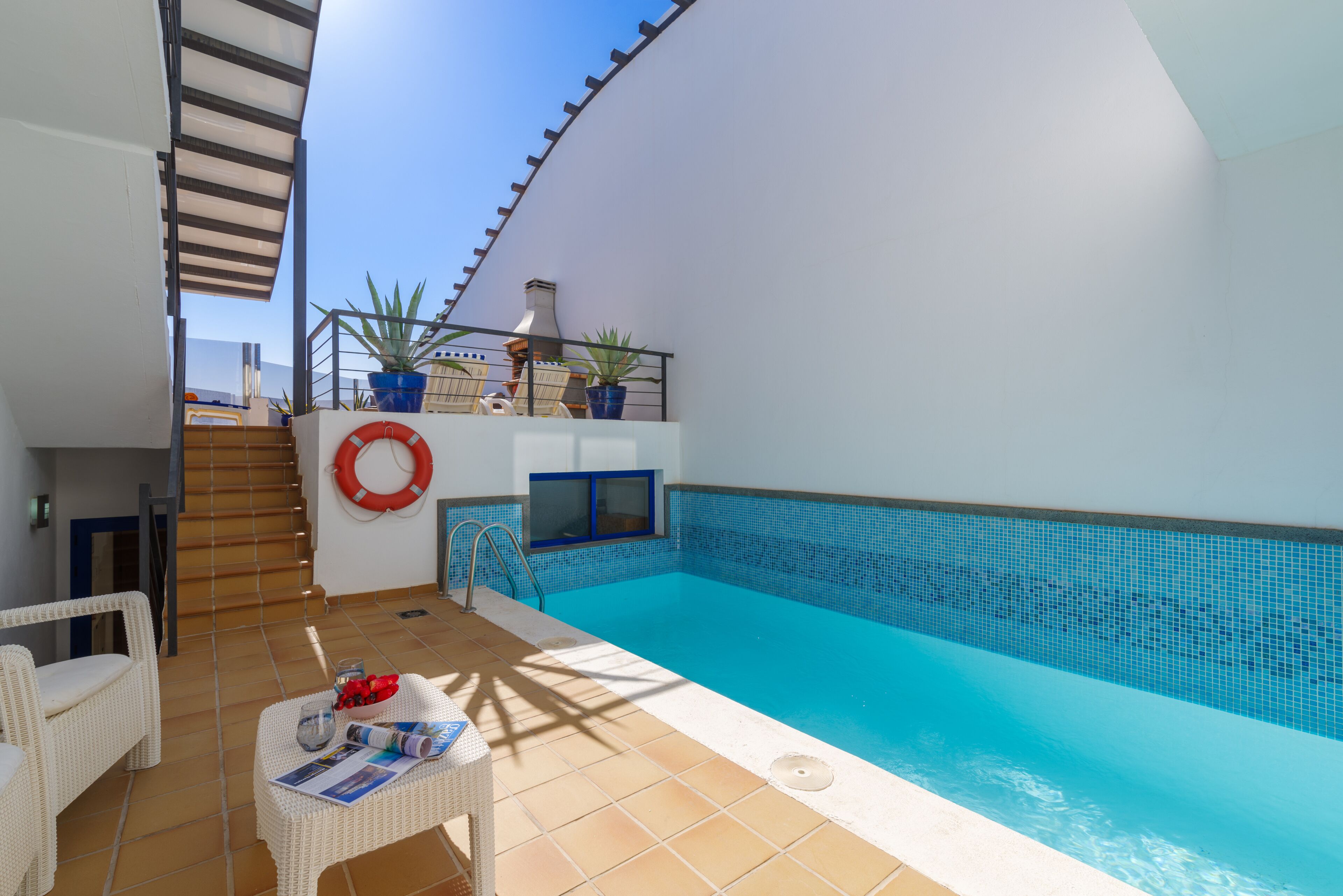 A modern outdoor pool area with a clear blue pool, white wicker furniture, and a staircase leading to a balcony with potted plants.