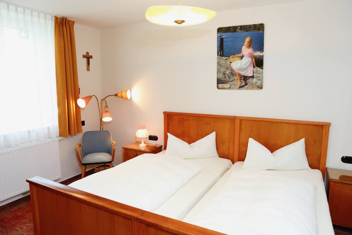 Een hotelkamer met een houten bed, een stoel, een nachtkastje met een lamp, een raam met gordijnen en een muur met een schilderij.