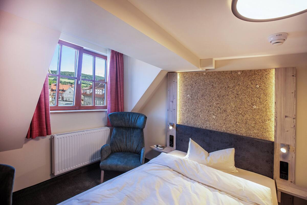 Een hotelkamer met een bed, een stoel, een raam met gordijnen, een radiator en een gestructureerde muur.