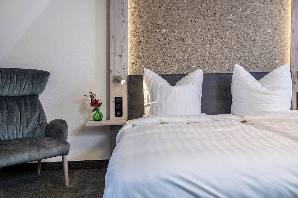 Een hotelkamer met een bed, een stoel, een nachtkastje en een wandplank met een bloevas.