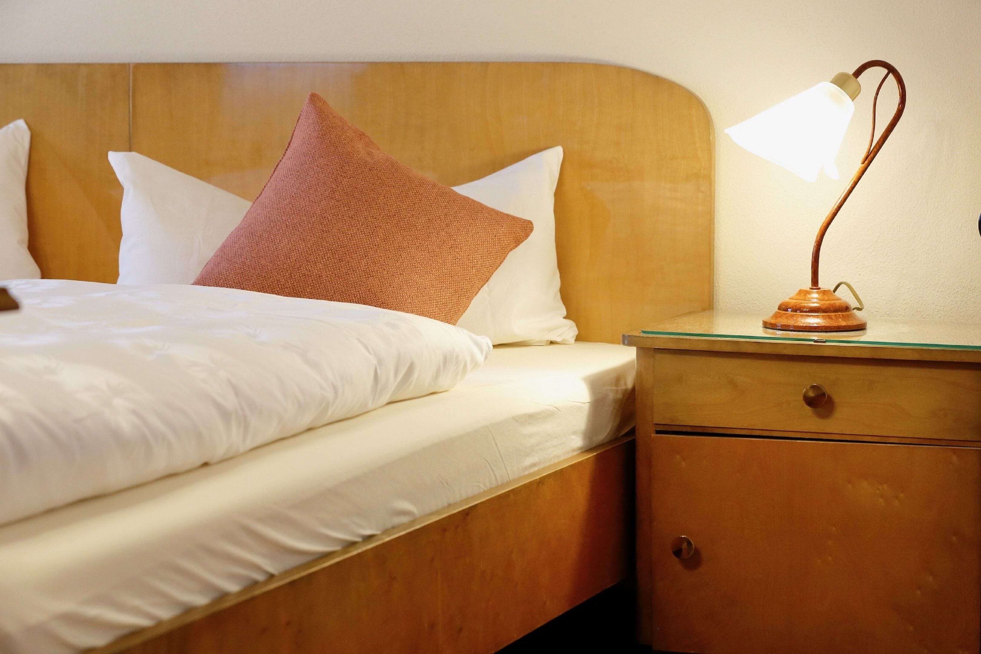 Een netjes opgemaakt bed met witte lakens, een houten hoofdbord, een bruin kussen en een nachtkastje met een lamp.