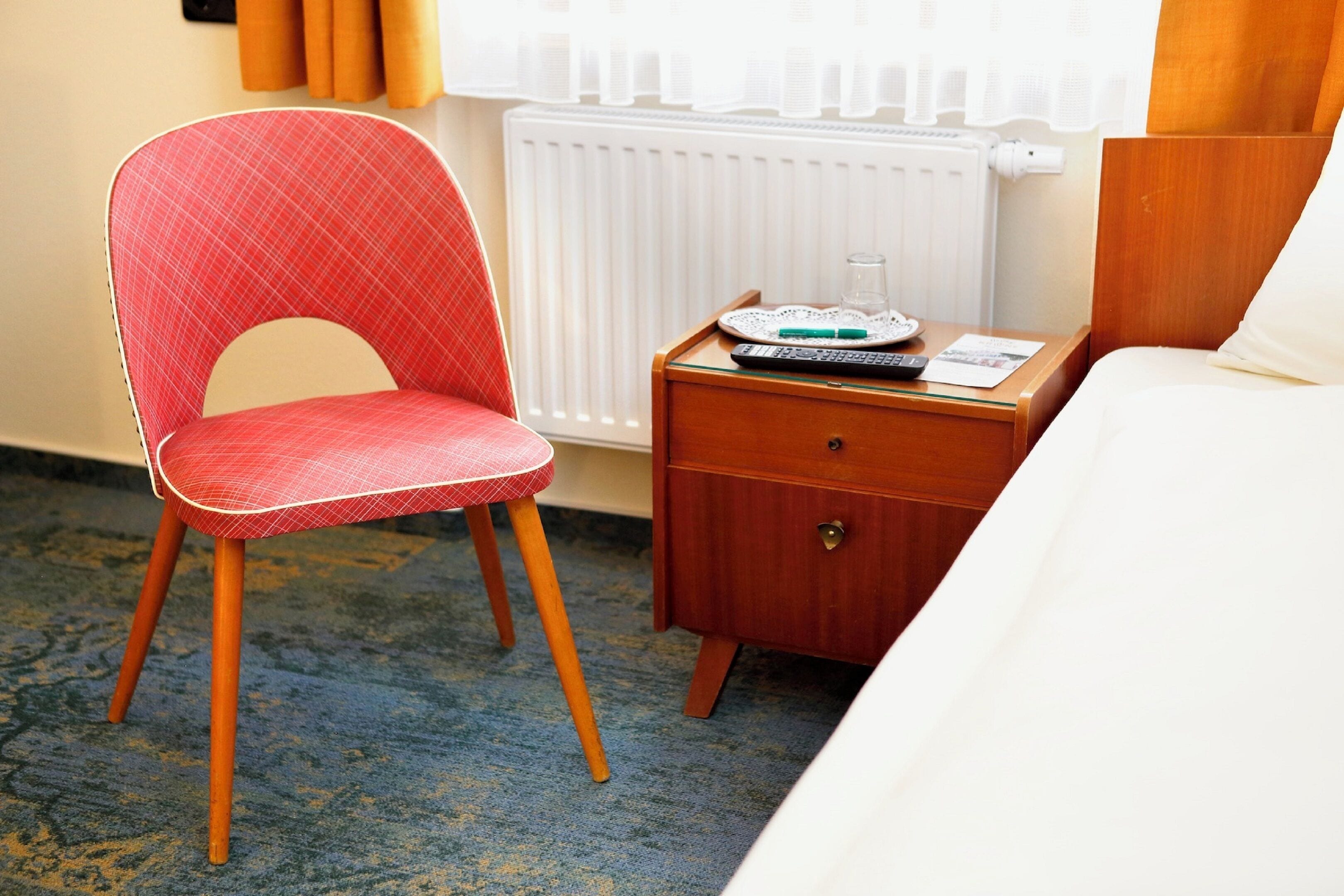 Una camera d'albergo con una poltrona imbottita rossa, un comodino in legno con un telecomando, un bicchiere d'acqua e un letto con biancheria da letto bianca.