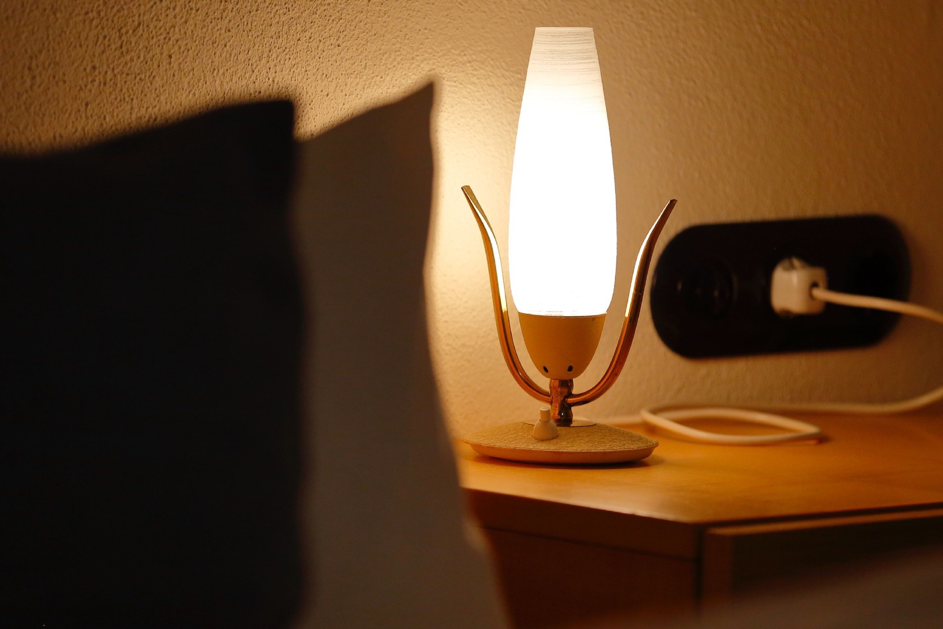 Eine Tischlampe mit weißem Schirm steht auf einem Holztisch, neben einer Steckdose und einem angeschlossenen Kabel.