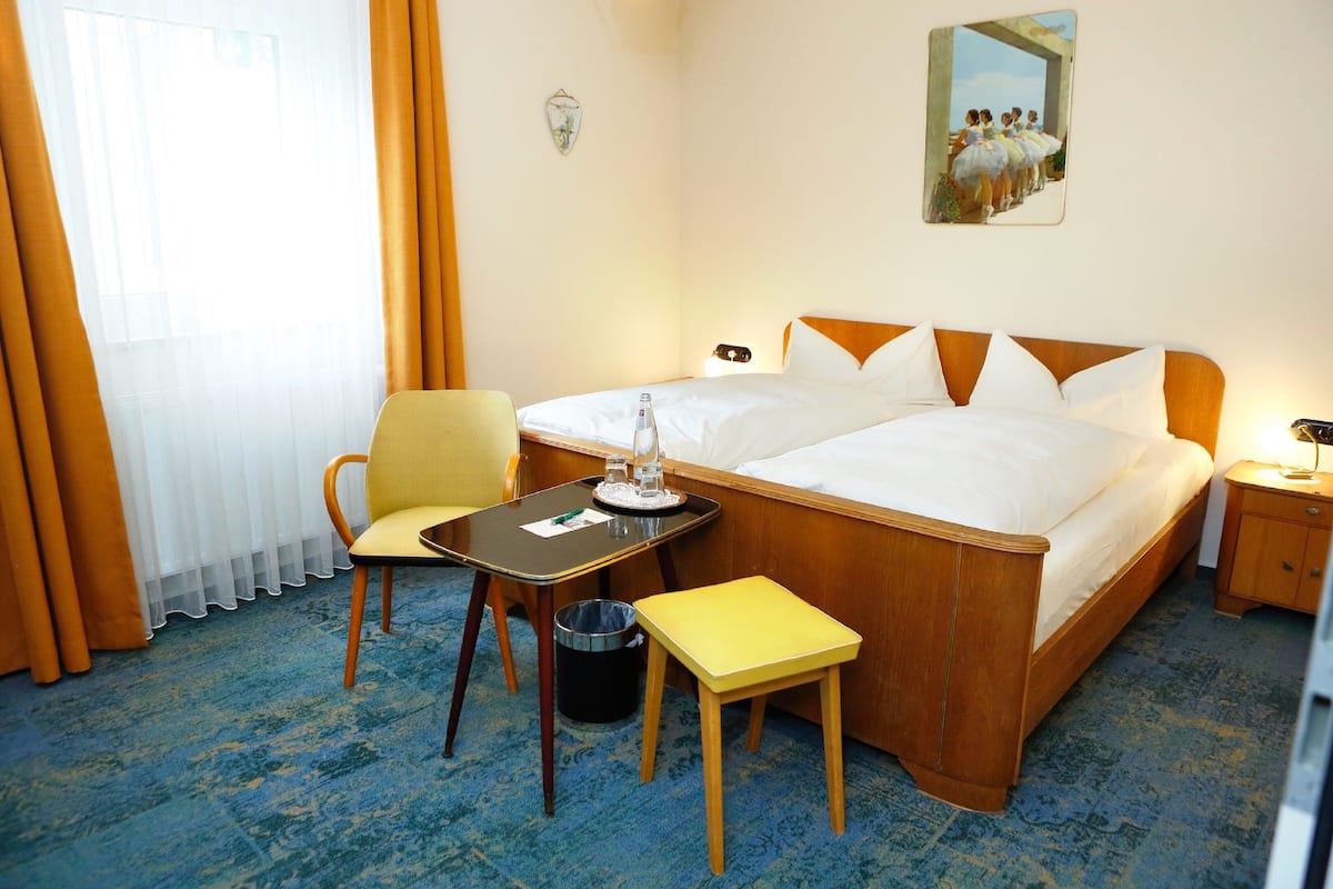 Hotelkamer met een tweepersoonsbed, een klein tafeltje met een stoel, een prullenbak en een schilderij aan de muur.