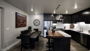A modern kitchen with dark cabinetry, a wooden island, stainless steel appliances, and a dining area with a dark table and chairs. - Sage Creek at Moab Tunnel Arch Hideaway (Moab)
