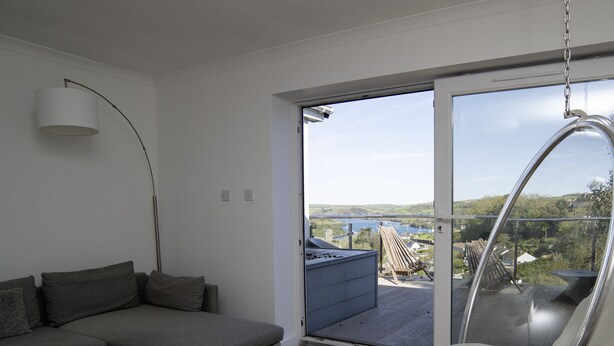 A modern living room with a grey sofa, a hanging chair, and a table with magazines. A large window offers a view of a balcony and a distant landscape. - Ultimate Coastal Escape | Stylish  | Estuary Views (Salcombe)
