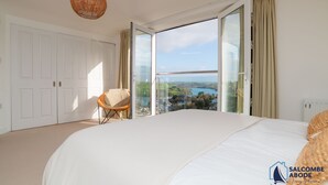 A bedroom with a large bed, a chair, a wicker ceiling light, and a view of a lake through a glass door. - Ultimate Coastal Escape | Stylish  | Estuary Views (Salcombe)