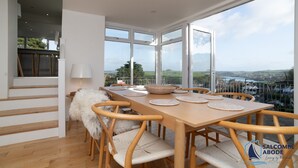 A dining area with a wooden table, woven placemats, and chairs. It features large windows with a scenic view. - Ultimate Coastal Escape | Stylish  | Estuary Views (Salcombe)