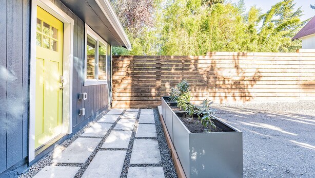 Una casa moderna con una puerta amarilla, una valla de madera y un jardín con plantas.