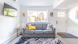 Una sala de estar moderna con un sofá gris, un tapete azul y blanco, y una ventana con vista a un edificio.