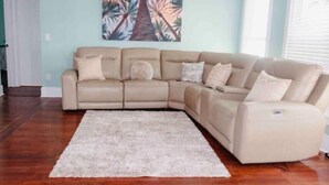 En beige hjørnesofa med mange puder, en hvid lurvet måtte og et maleri af en tropisk palme på væggen.