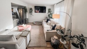 A modern living room with a sofa, a coffee table, and a TV. There is a kitchen area in the background with a fridge and cabinets.