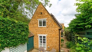 A stone house with a triangular roof, a small garden, and a table with plates and a glass of juice.