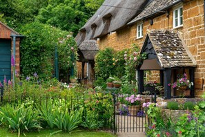 Une maison de campagne traditionnelle en pierre, avec un toit de chaume, un petit jardin agrémenté de diverses plantes et fleurs, et une pancarte indiquant « MAISON DE MARTIN ».