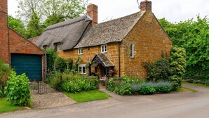A stone house with a thatched roof, a green garage, and a well-maintained garden with various plants and flowers.