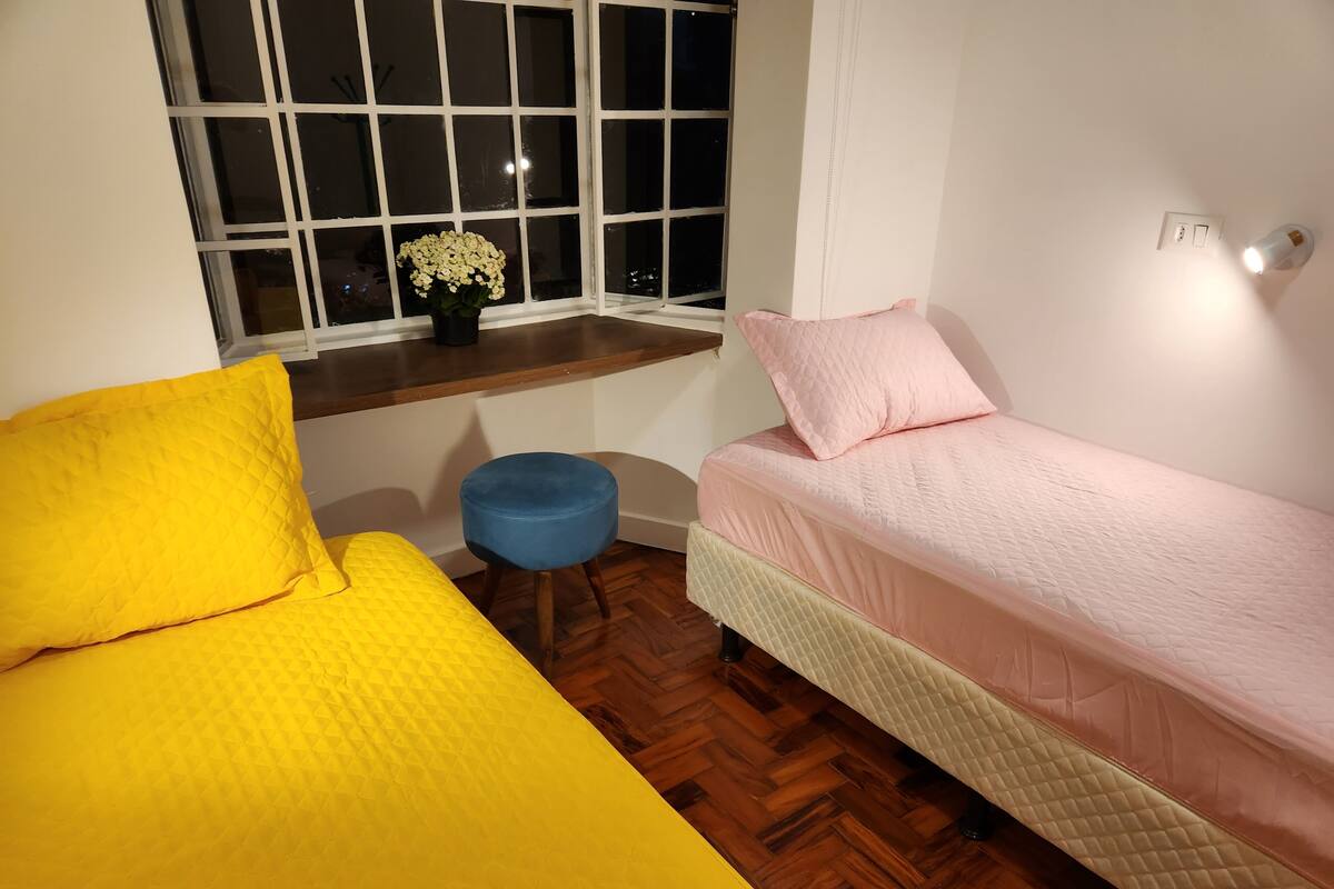 A room with two beds, one with yellow bedding and the other with pink bedding, a small stool, a window with a view of the night sky, and a wall socket.